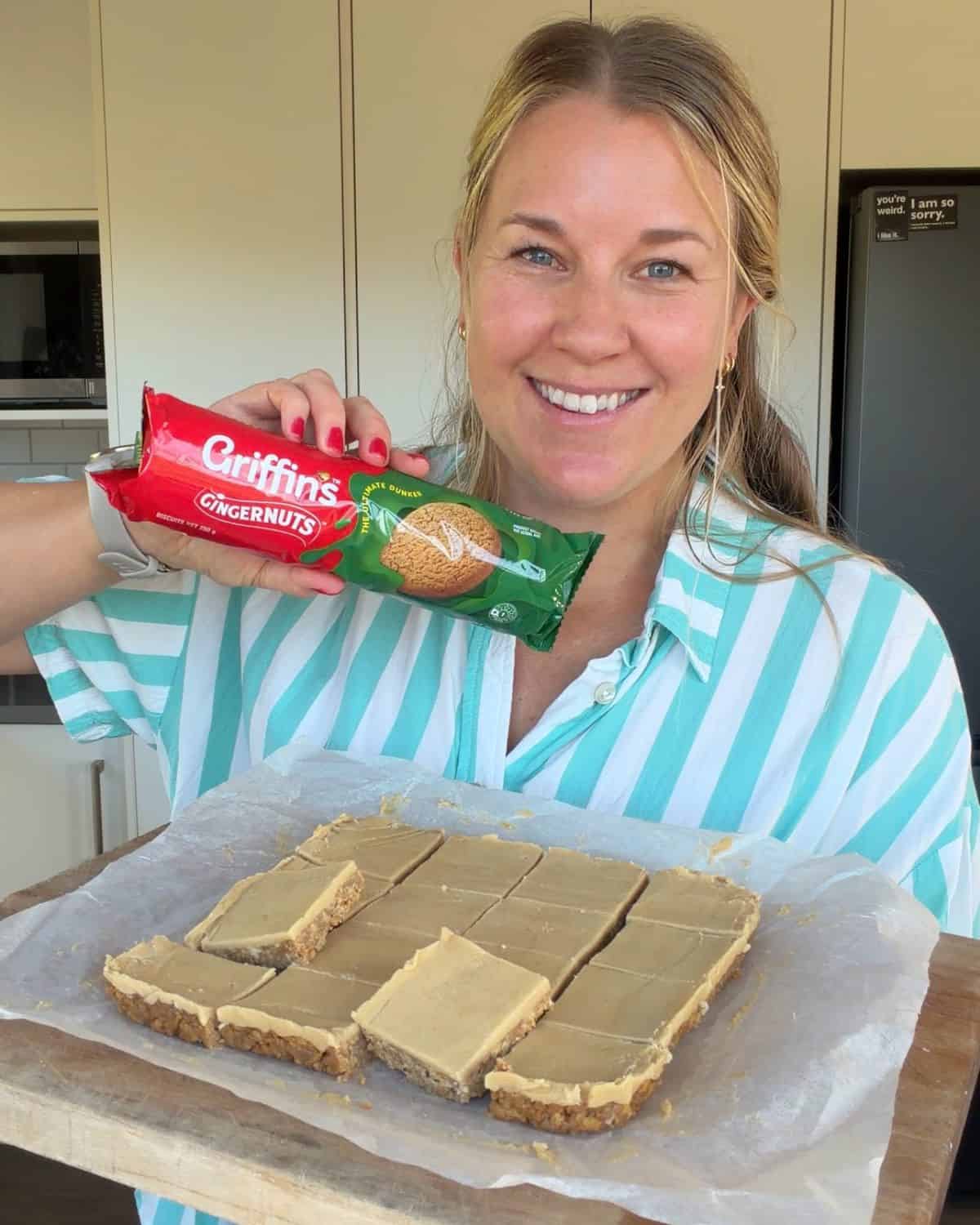 A woman in a striped shirt holds a packet of Griffins Gingernuts biscuits and stands behind a tray of sliced dessert bars on parchment paper.