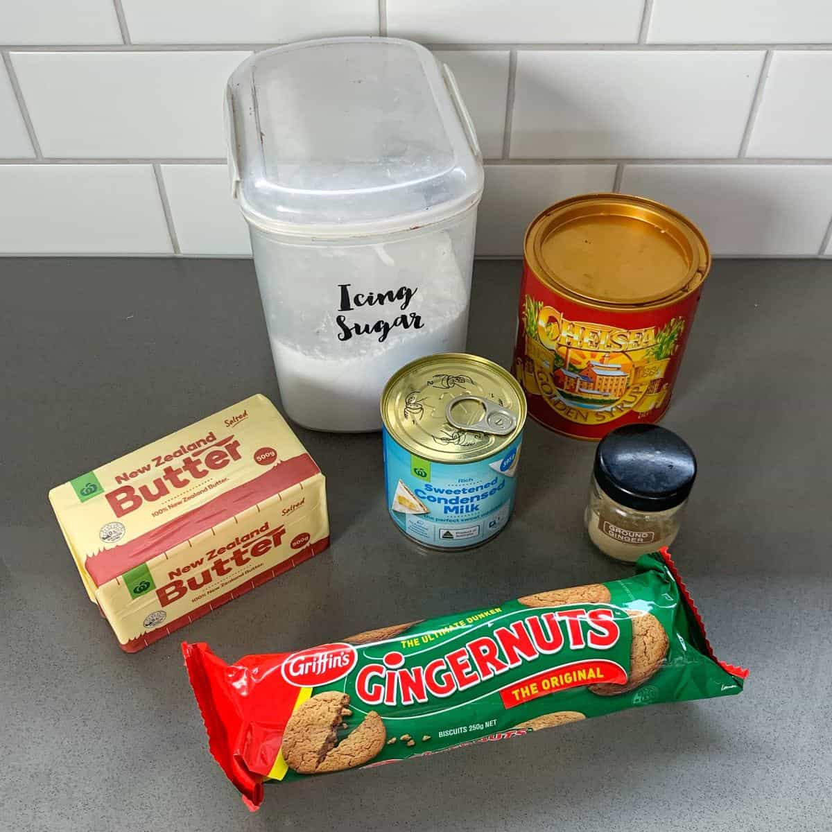 A collection of baking ingredients on a counter: icing sugar, butter, condensed milk, gingersnap biscuits, ground ginger, and a tin of crystallized ginger.