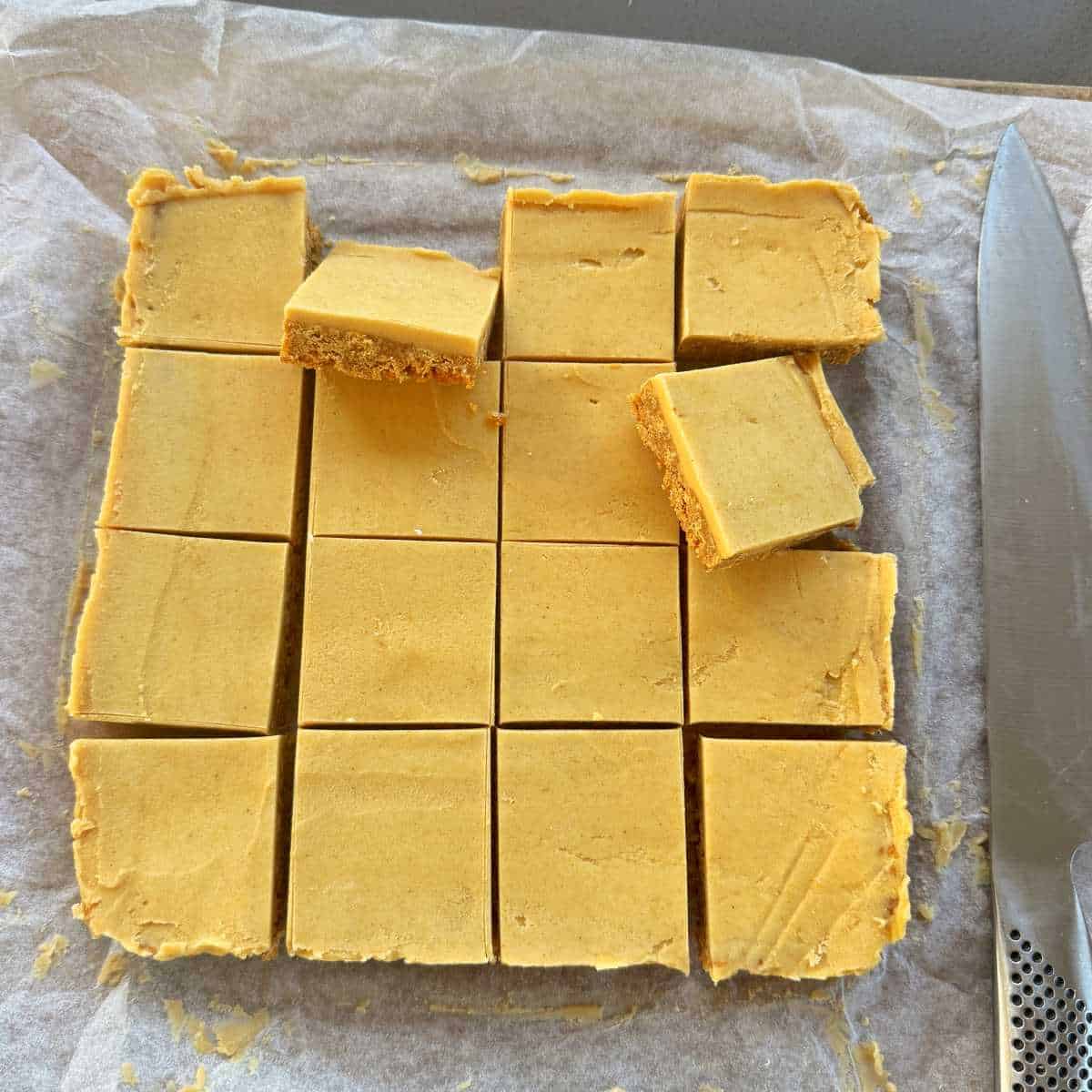No-Bake Gingernut Slice A tray of fifteen square pieces of fudge on parchment paper, with a large knife beside them and two pieces stacked on top.