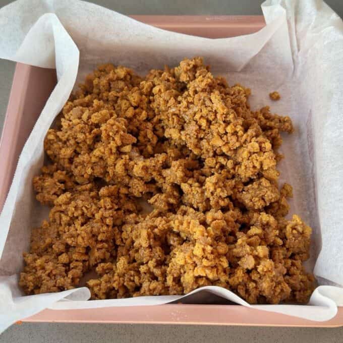 No-Bake Gingernut Slice Crumble mixture spread in a parchment-lined square baking pan, ready for the next step in a recipe.