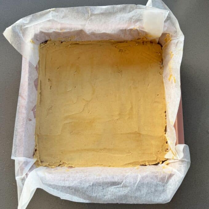No-Bake Gingernut Slice A square baking pan lined with parchment paper, containing an unbaked or freshly baked flat layer of light brown dough or batter.
