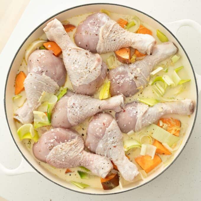 One Pot Chicken, Leek and Orzo Bake Raw chicken drumsticks arranged in a white dish with chopped sweet potatoes and leeks, seasoned and ready to be cooked in a creamy liquid.