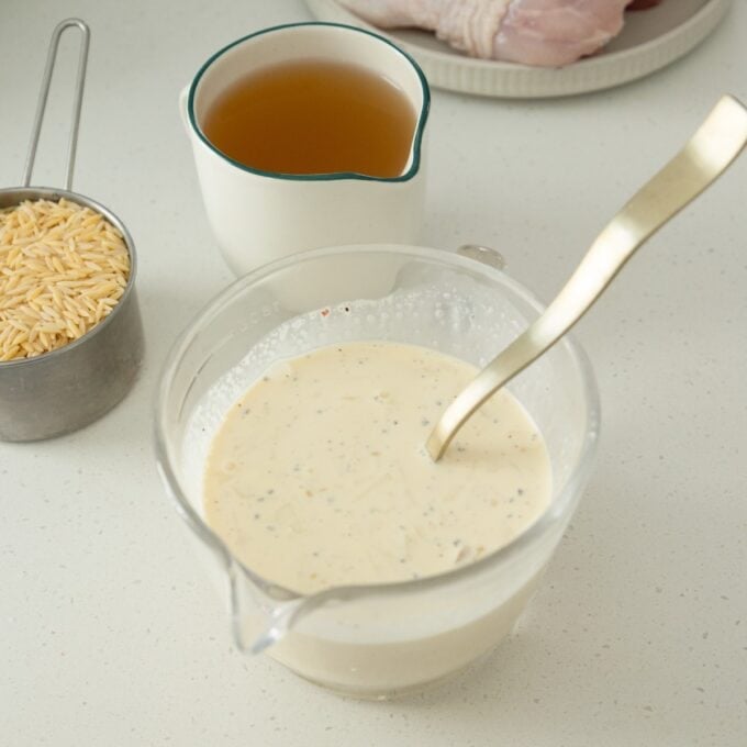 One Pot Chicken, Leek and Orzo Bake Measuring cup with creamy sauce and spoon, metal cup of dry orzo, a cup of broth, and raw chicken on a plate, all on a light counter.