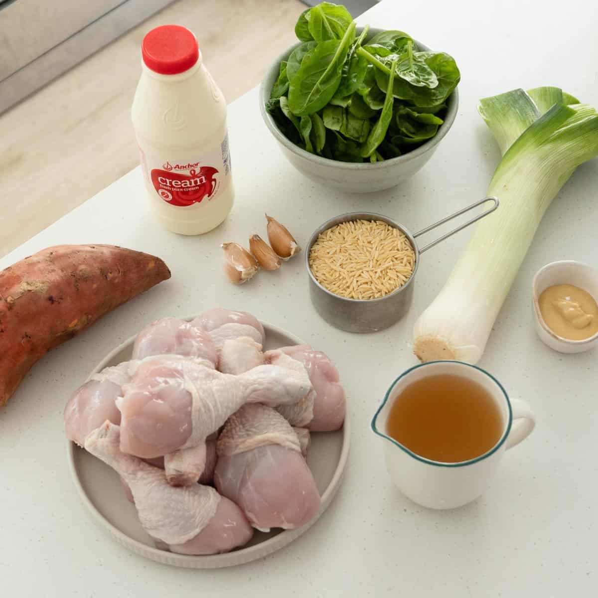 One Pot Chicken, Leek and Orzo Bake Raw chicken drumsticks, sweet potato, heavy cream, garlic cloves, baby spinach, orzo pasta, leek, Dijon mustard, and chicken broth arranged on a white countertop.
