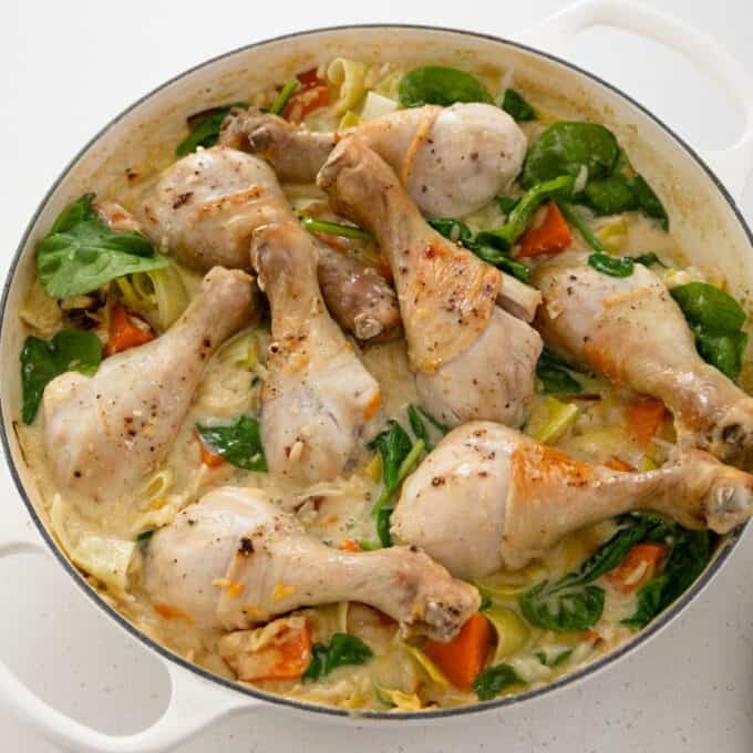 One Pot Chicken, Leek and Orzo Bake Chicken drumsticks cooked with spinach, chopped vegetables, and a creamy sauce in a white, round dish on a white surface.