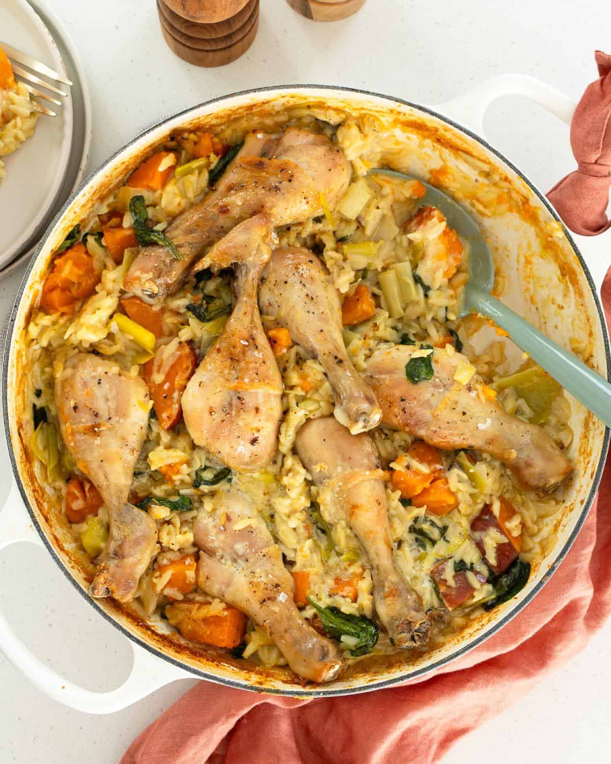 One Pot Chicken, Leek and Orzo Bake Baked chicken drumsticks on top of creamy rice with vegetables in a white casserole dish, served with a blue spoon.
