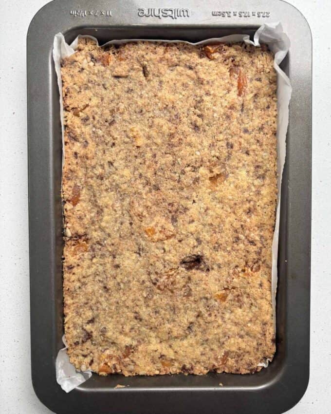 Gluten Free Bumper Bars A rectangular baking tray filled with a baked oat and dried fruit mixture, lined with parchment paper, sits on a white surface.