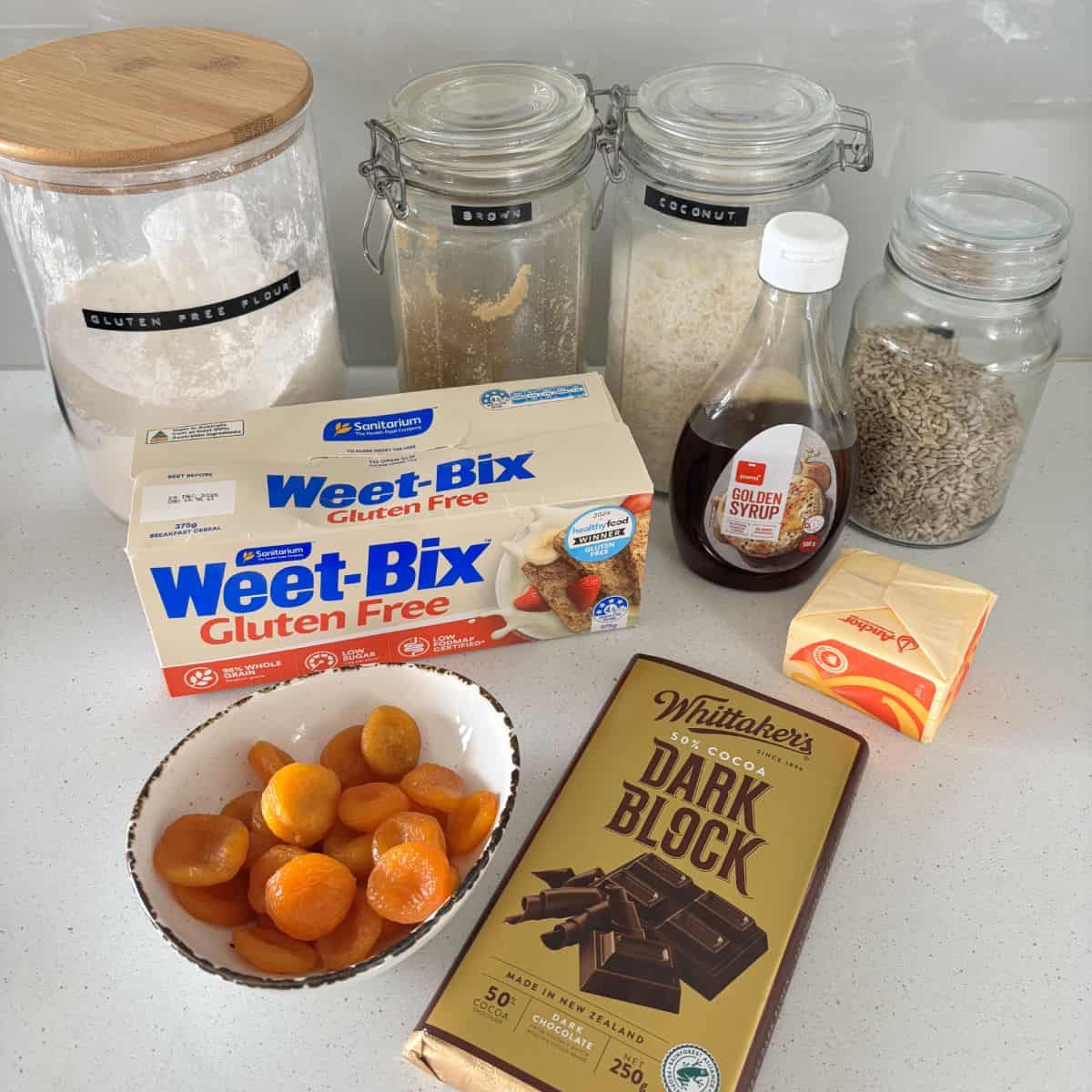 Gluten Free Bumper Bars Various baking ingredients are arranged on a countertop, including jars of flour and coconut, Weet-Bix, sunflower seeds, golden syrup, butter, dried apricots, and a bar of dark chocolate.