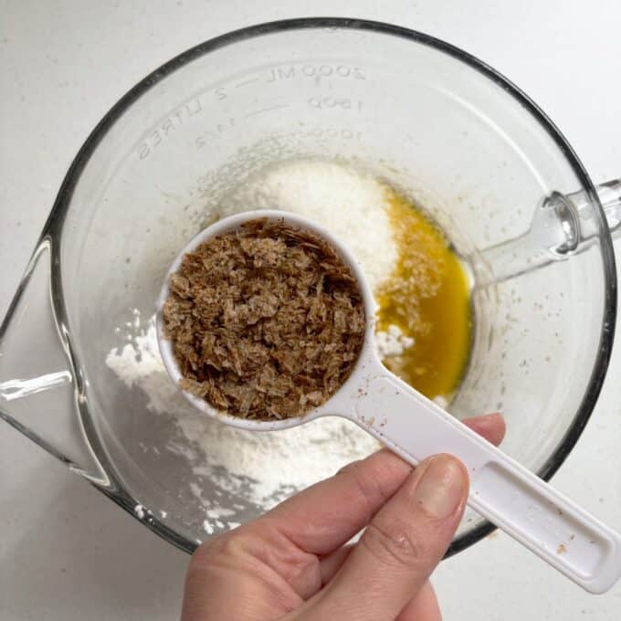 Gluten Free Bumper Bars A hand holds a white measuring cup of bran flakes over a glass bowl containing flour and melted butter on a white surface.