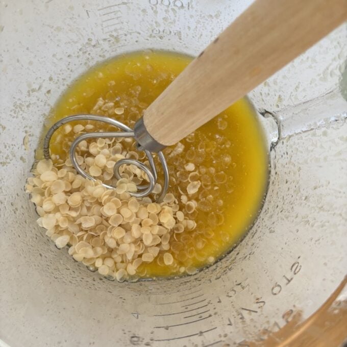Gluten Free Bumper Bars A glass measuring cup with melted yellow liquid, solid wax pellets, and a metal whisk with a wooden handle inside.