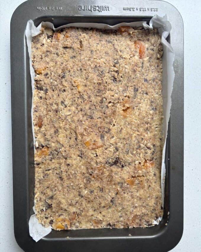Gluten Free Bumper Bars A rectangular baking tray lined with parchment paper, filled with an unbaked mixture containing nuts, oats, and dried fruit.