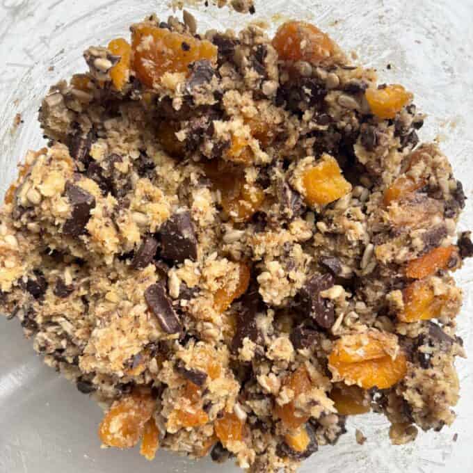 Gluten Free Bumper Bars Cookie dough mixture with visible chunks of dark chocolate, dried apricots, sunflower seeds, oats, and other ingredients in a glass bowl.