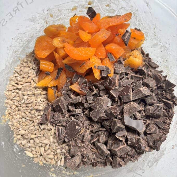 Gluten Free Bumper Bars Chopped dried apricots, sunflower seeds, and dark chocolate chunks on top of dry ingredients in a mixing bowl.