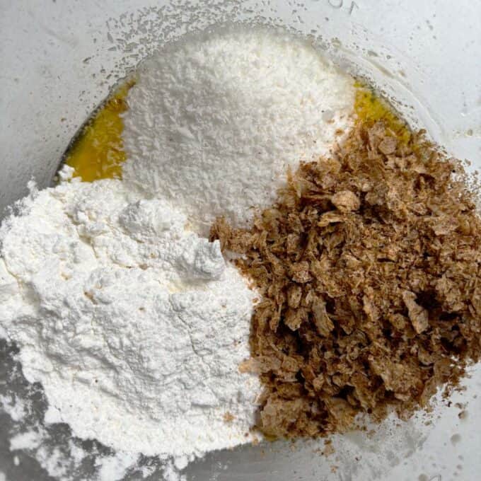 Gluten Free Bumper Bars A mixing bowl containing shredded coconut, flour, crushed bran flakes, and a yellow liquid ingredient, possibly melted butter or egg.