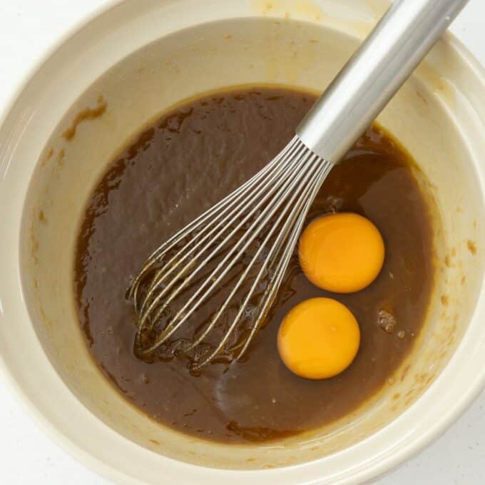 Hot Cross Blondies A mixing bowl with brown batter, two raw eggs, and a metal whisk partially submerged in the mixture.
