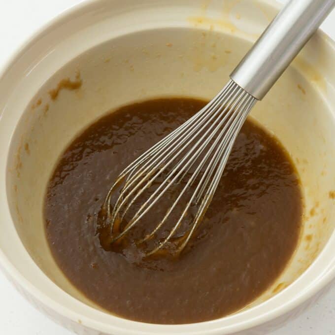 Hot Cross Blondies A metal whisk in a beige bowl containing a thick brown mixture, possibly batter or sauce, on a white surface.