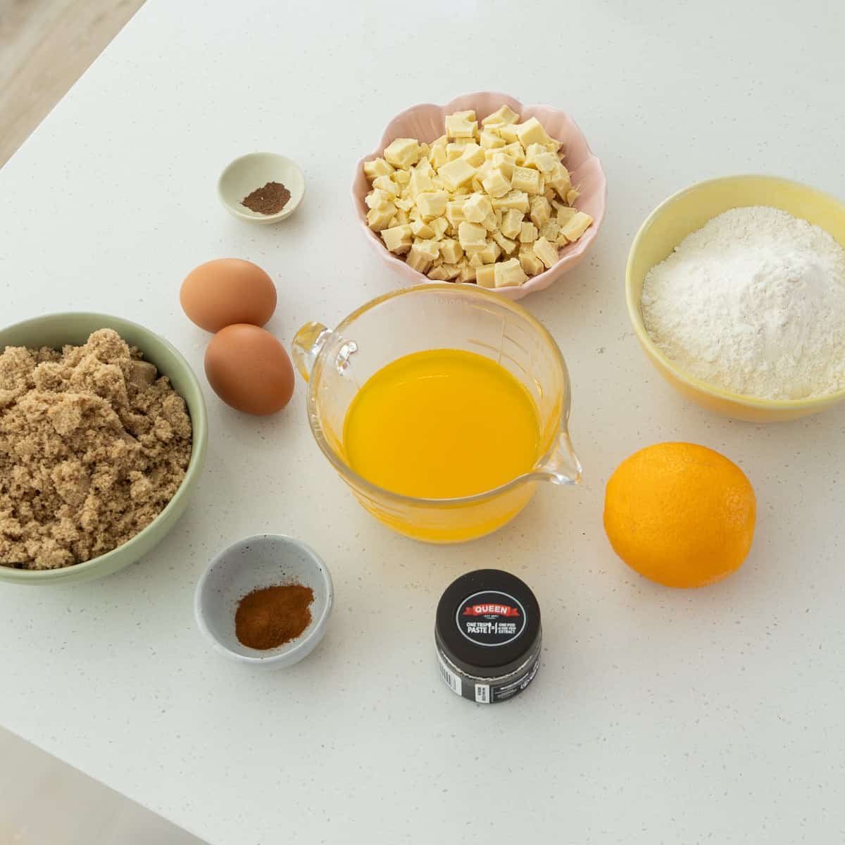 Baking ingredients on a white countertop, including eggs, brown sugar, melted butter, flour, orange, white chocolate chunks, cinnamon, nutmeg, and a small jar of ground cinnamon.