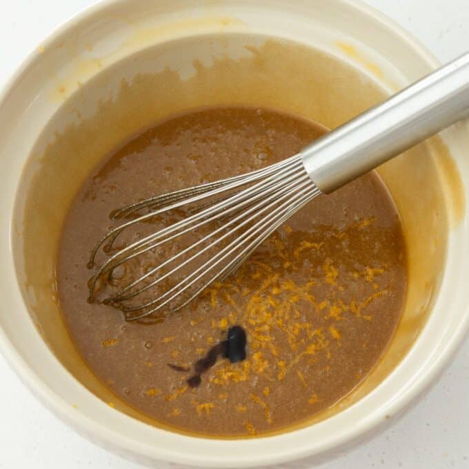 Hot Cross Blondies A whisk rests in a bowl filled with brown batter, with some grated orange zest and a drop of dark liquid visible on the surface.