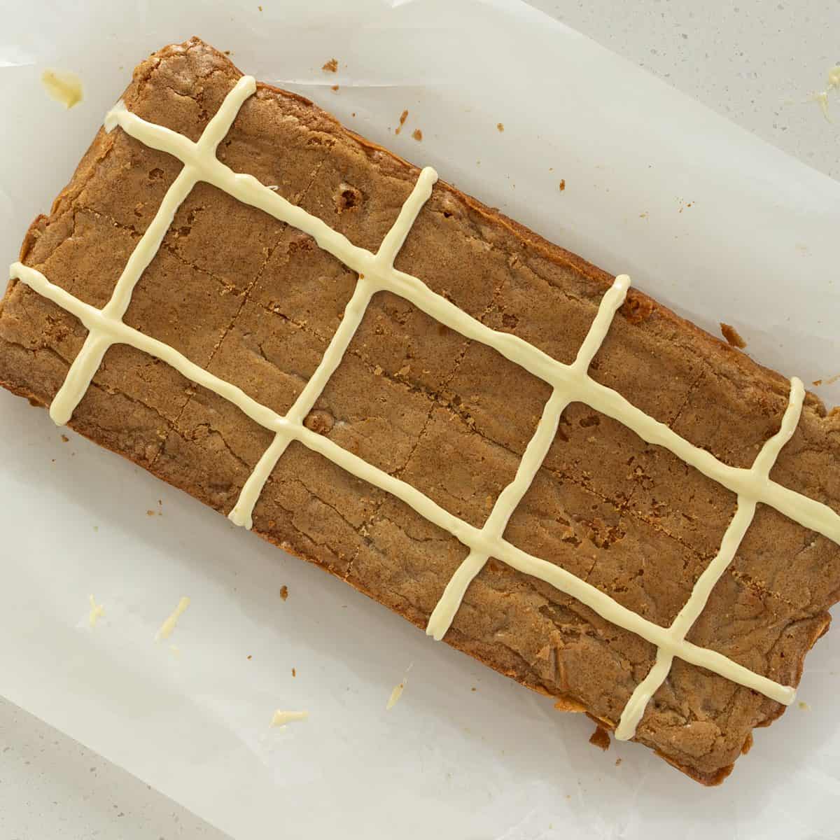 Hot Cross Blondies A rectangular blondie with a light brown top is decorated with white icing in a grid pattern, sitting on parchment paper.