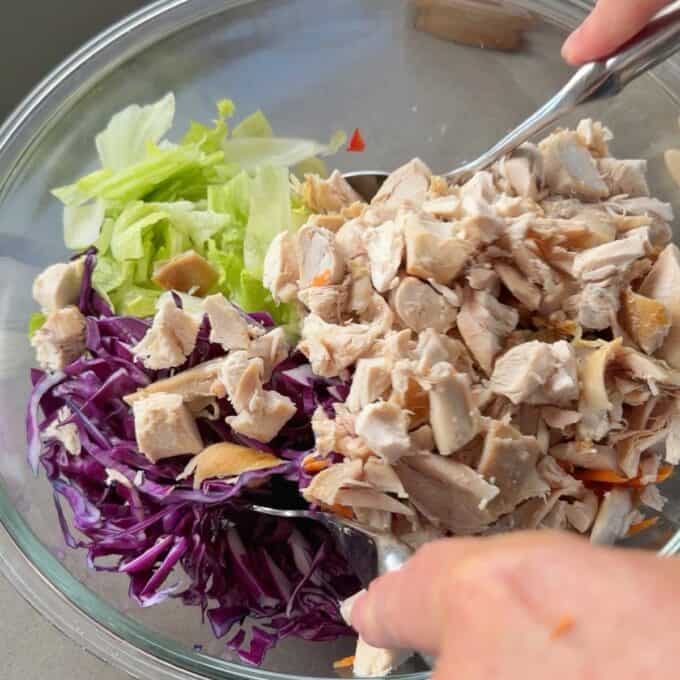 Rotisserie Chicken Salad A glass bowl containing chopped lettuce, shredded purple cabbage, and cooked diced chicken being mixed with a spoon.