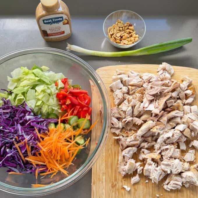 Rotisserie Chicken Salad Chopped cooked chicken on a cutting board next to a bowl of lettuce, red bell pepper, purple cabbage, carrots, and cucumbers, with peanuts, a green onion, and sauce nearby.