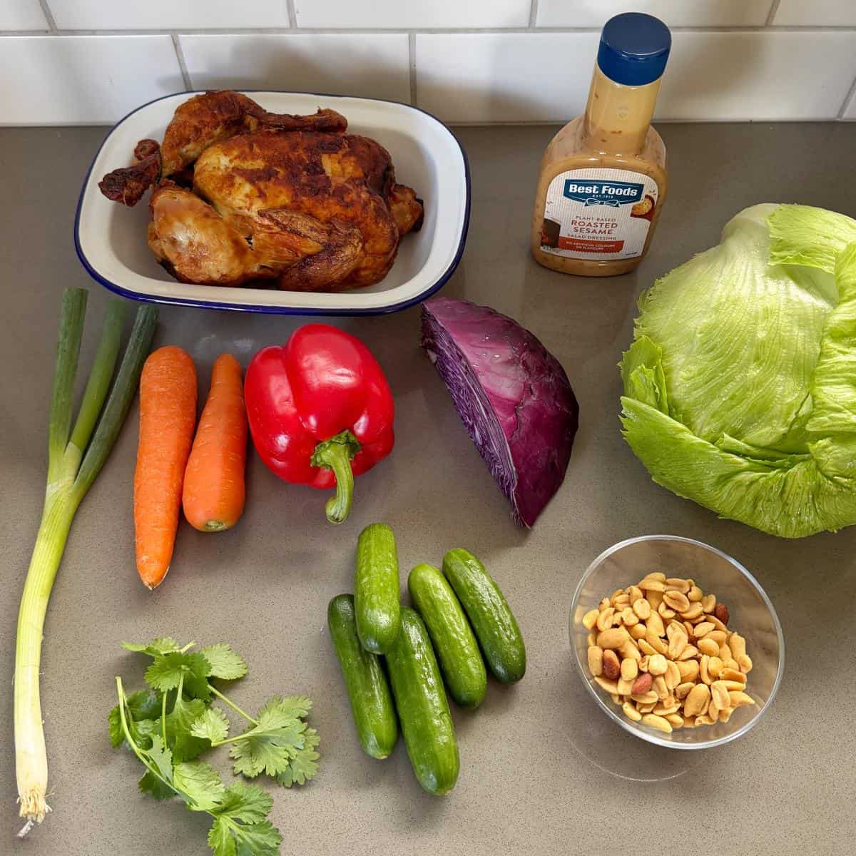 Rotisserie Chicken Salad A countertop with a roast chicken, lettuce, red cabbage, carrots, red bell pepper, green onions, cucumbers, cilantro, mixed nuts, and a bottle of salad dressing.