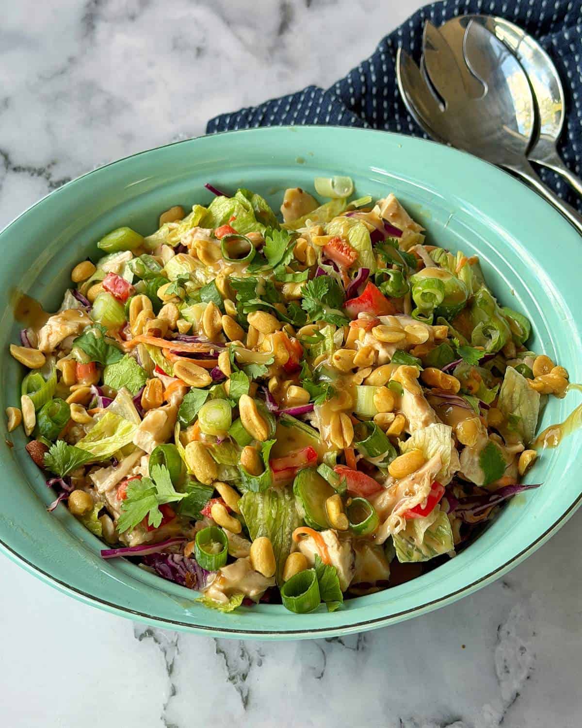 Rotisserie Chicken Salad A bowl of chopped salad with lettuce, peanuts, green onions, cilantro, and shredded vegetables on a marble surface, next to serving utensils and a napkin.
