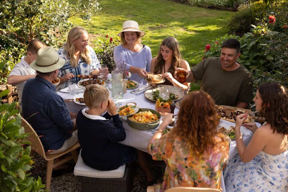 Eight people gather around an outdoor table in a garden, sharing a meal and smiling. Various dishes, drinks, and flowers are set on the table. Lush greenery surrounds the scene.