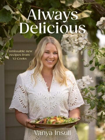 Woman in a white dress holding a platter of food stands outdoors under flowering branches. Text reads: Always Delicious, Unmissable new recipes from VJ Cooks, Vanya Insull..