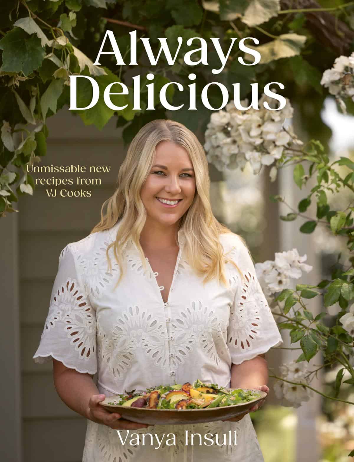 Woman in a white dress holding a platter of food stands outdoors under flowering branches. Text reads: Always Delicious, Unmissable new recipes from VJ Cooks, Vanya Insull..