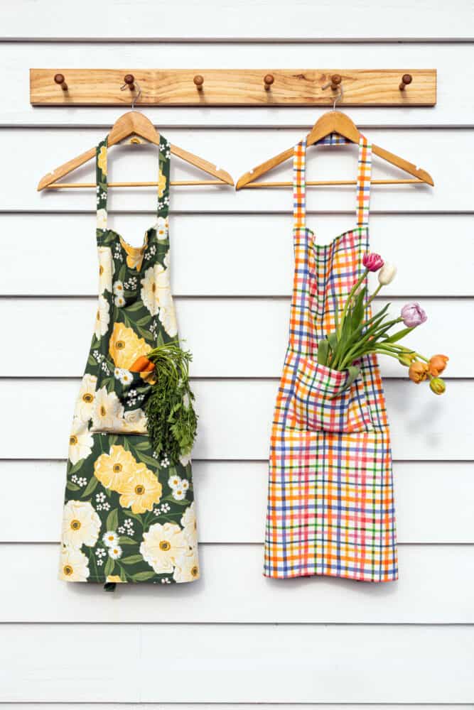 Two aprons on wooden hangers hang from hooks on a white wall; the left apron is floral with carrots in its pocket, the right is plaid with tulips in its pocket.
