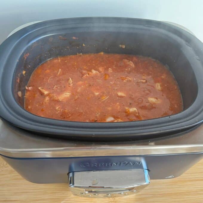 Slow Cooker Chicken Ragu A slow cooker filled with a tomato-based stew containing chunks of meat and vegetables sits on a wooden surface.