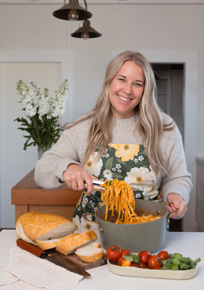 Woman in a floral apron serves spaghetti from a pot, with sliced bread and fresh vegetables on the counter in a bright kitchen.