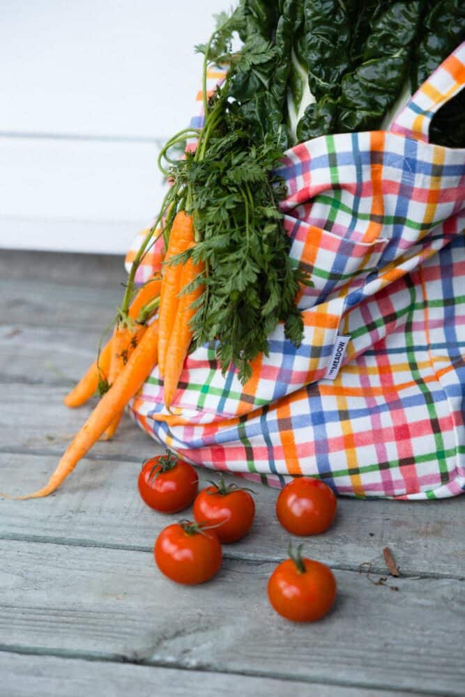A plaid cloth bag with leafy greens and carrots, with several tomatoes on a wooden surface.
