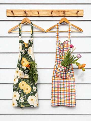 Two aprons hang on wooden hooks; the left apron has a floral pattern with carrots in its pocket, and the right apron has a plaid pattern with tulips in its pocket.