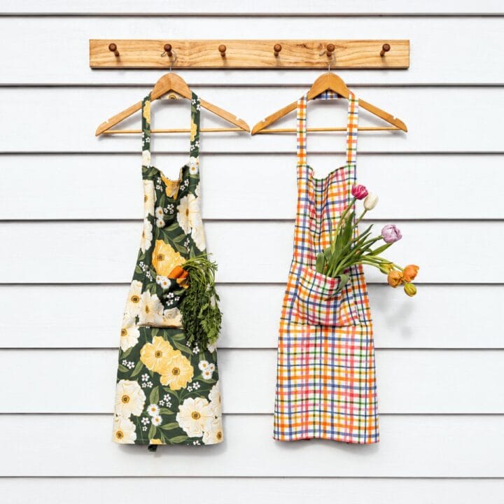 Two aprons hang on wooden hooks; the left apron has a floral pattern with carrots in its pocket, and the right apron has a plaid pattern with tulips in its pocket.