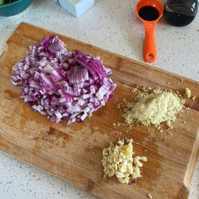 Chicken Larb Chopped red onion, minced garlic, and grated ginger on a wooden cutting board with a measuring spoon and soy sauce nearby.