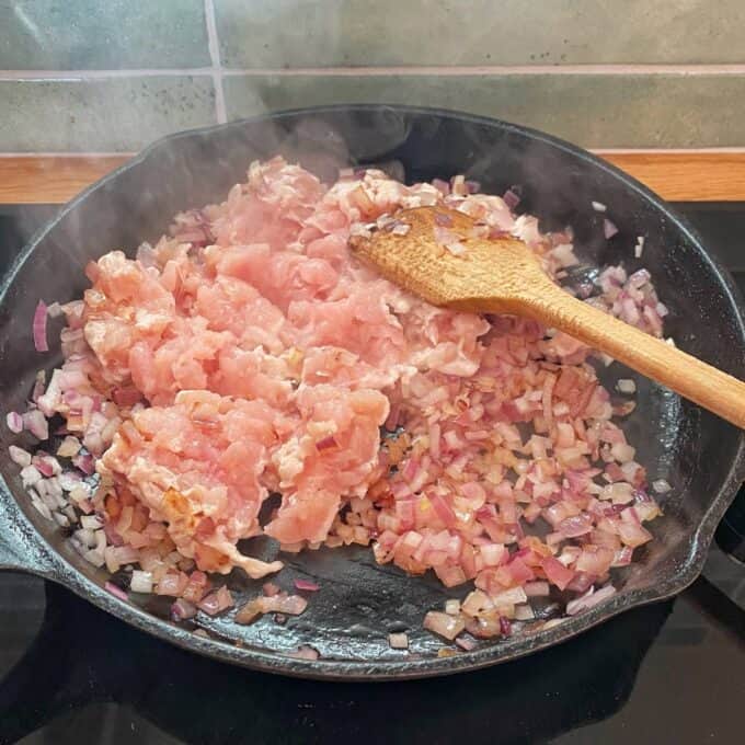 Chicken Larb Chopped raw chicken and diced onions are being cooked in a cast iron skillet, stirred with a wooden spoon. Steam rises from the pan.