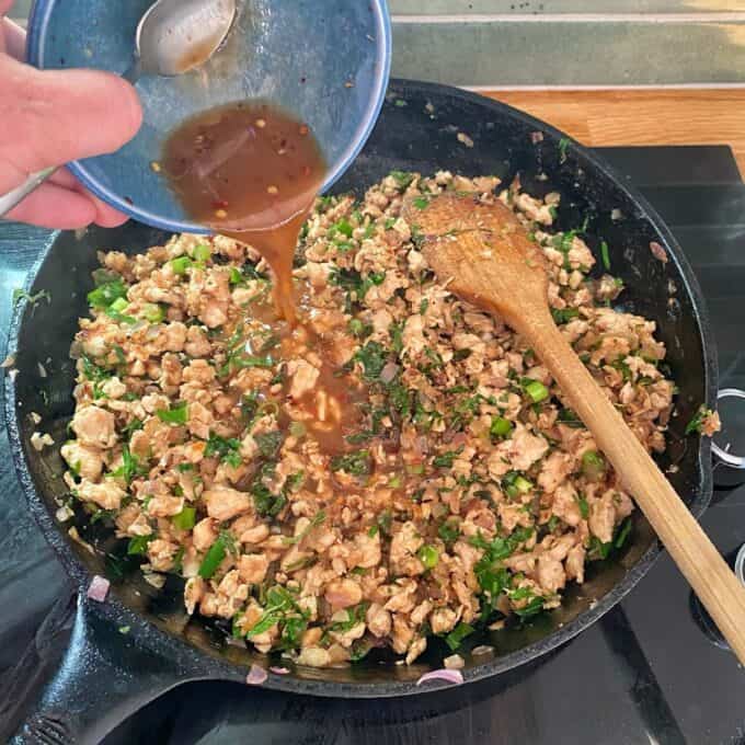 Chicken Larb A hand pours sauce from a small bowl into a skillet filled with cooked ground meat and chopped green herbs; a wooden spoon rests inside the skillet.