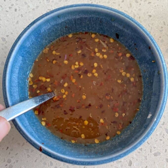 Chicken Larb A blue bowl with a metal spoon contains a brown liquid sauce mixed with chili flakes, placed on a light speckled countertop.