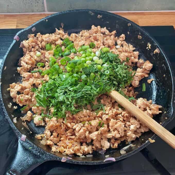 Chicken Larb Ground meat cooks in a cast iron skillet with chopped green onions and herbs being stirred in with a wooden spoon.