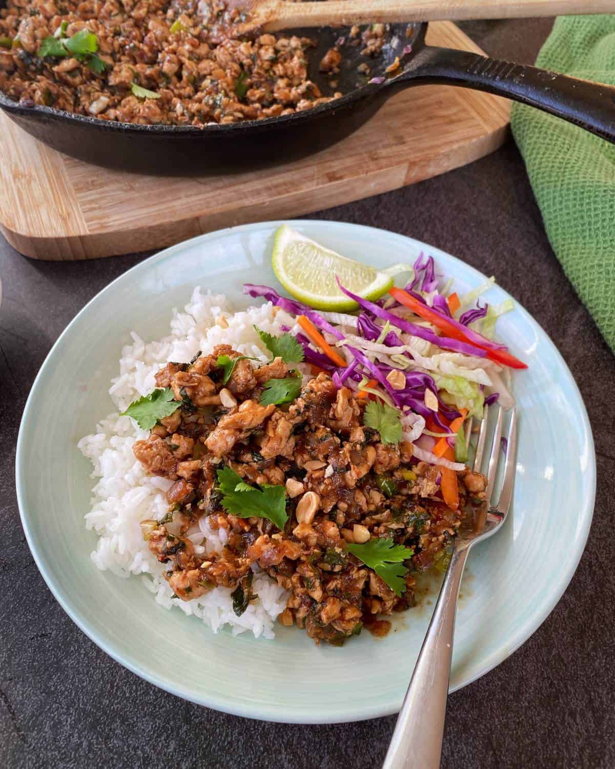 Chicken Larb A plate of white rice topped with minced meat, chopped herbs, and peanuts, served with sliced lime, shredded cabbage, and carrot, with a fork on the side. A skillet is in the background.