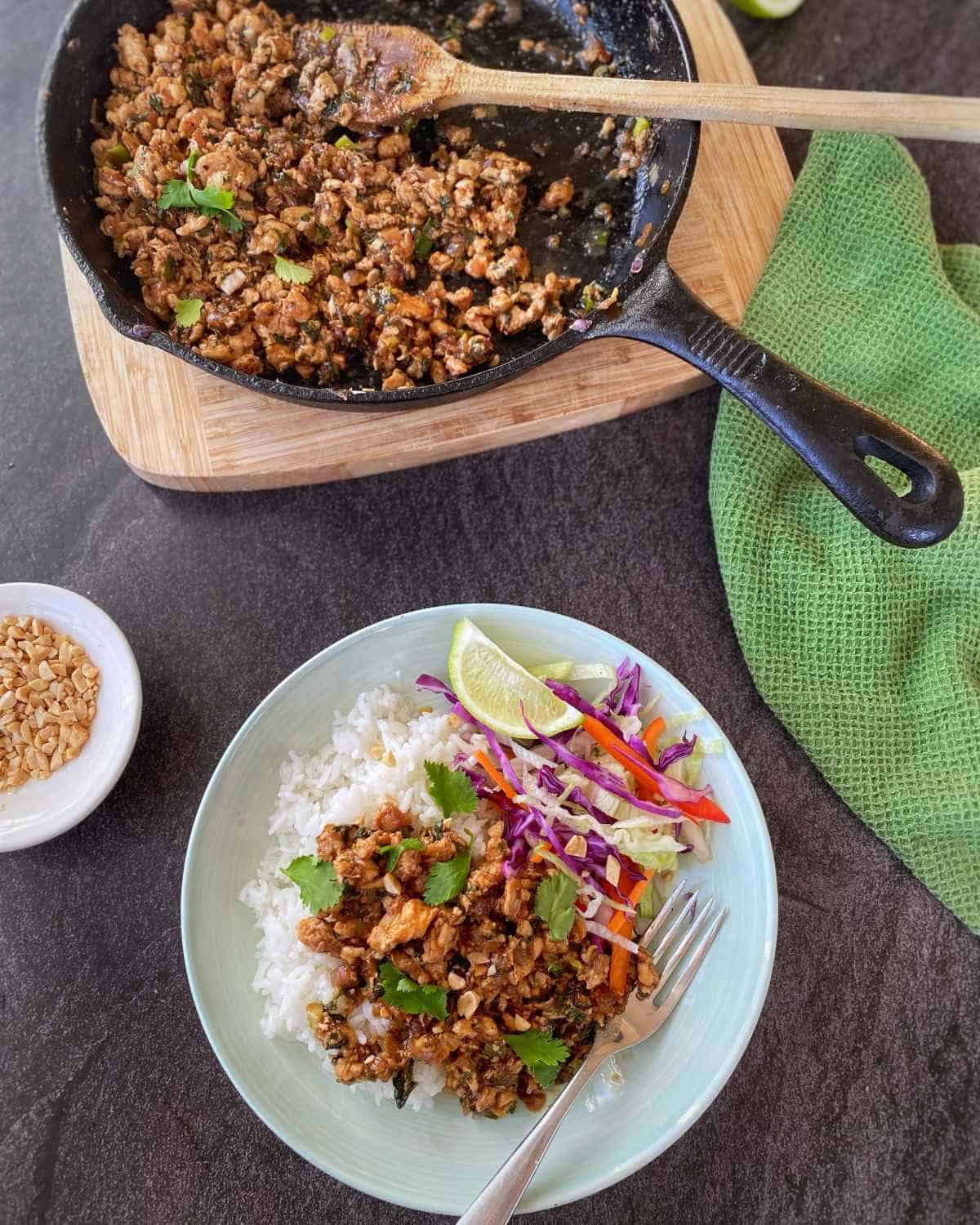 Chicken Larb A plate of rice with ground meat, vegetables, lime wedges, and herbs next to a skillet of cooked meat, a small bowl of peanuts, and a green cloth on a dark surface.