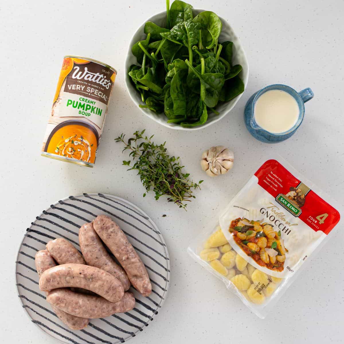 Creamy Pumpkin and Sausage Gnocchi A can of pumpkin soup, fresh spinach, cream, a garlic bulb, thyme, sausages, and a package of gnocchi arranged on a white surface.