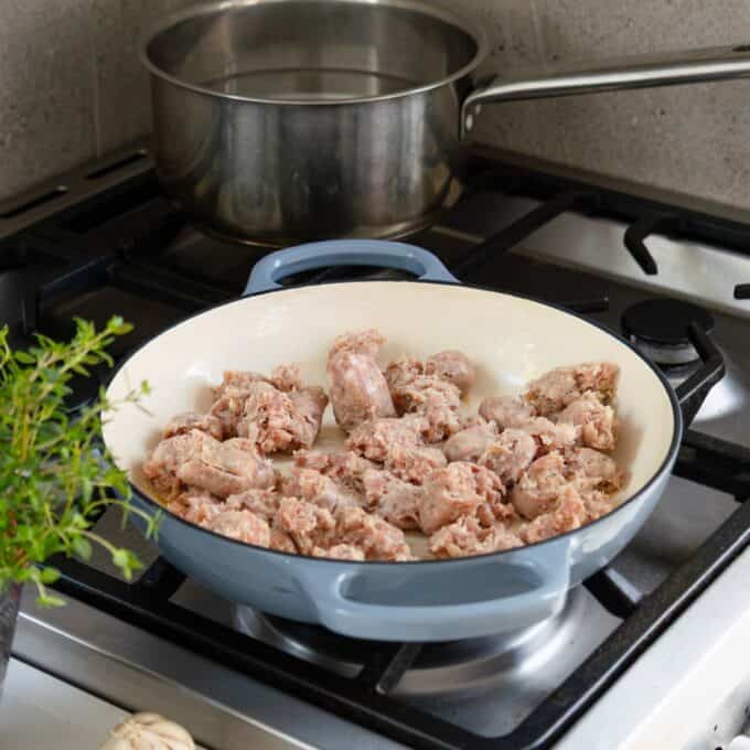 Creamy Pumpkin and Sausage Gnocchi Chunks of raw sausage are cooking in a blue skillet on a stove, with a pot of water simmering on a nearby burner.