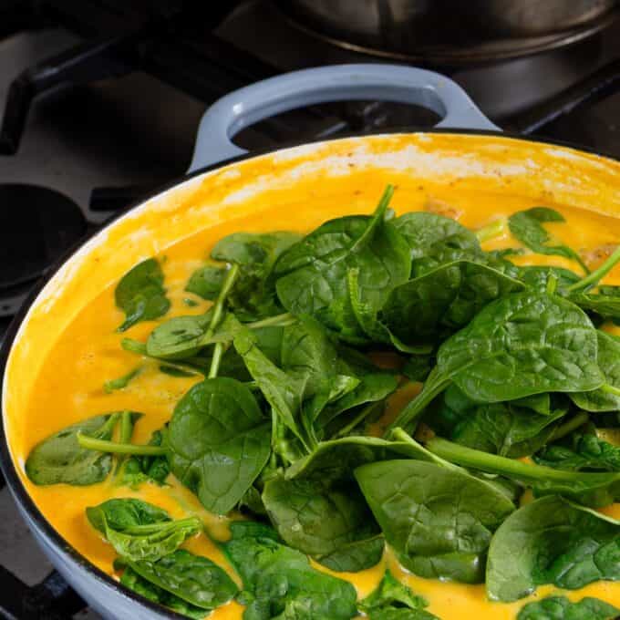 Creamy Pumpkin and Sausage Gnocchi A skillet on a stove contains a yellow sauce with fresh spinach leaves piled on top, ready to be cooked.