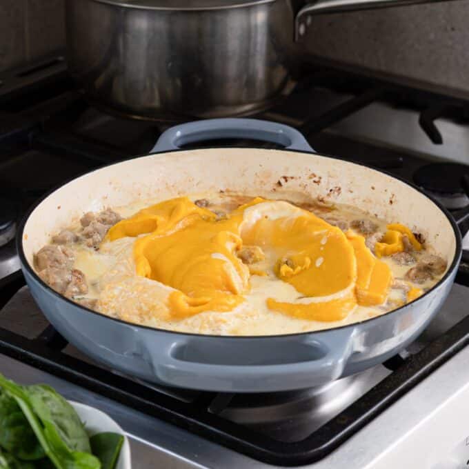 Creamy Pumpkin and Sausage Gnocchi A blue pan on a stove contains a creamy mixture with pieces of cooked squash and mushrooms. A stainless steel pot sits on the burner behind it.