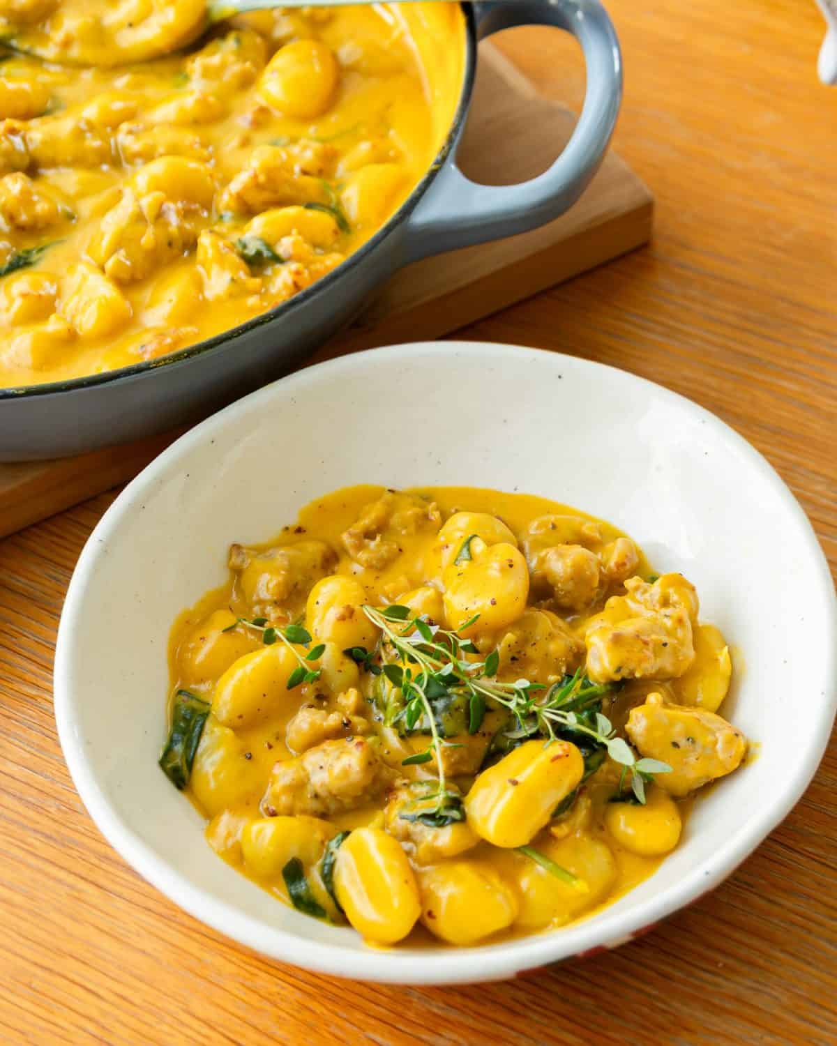 Creamy Pumpkin and Sausage Gnocchi A bowl of gnocchi in a creamy yellow sauce with herbs, alongside a pan containing more of the same dish, on a wooden table.
