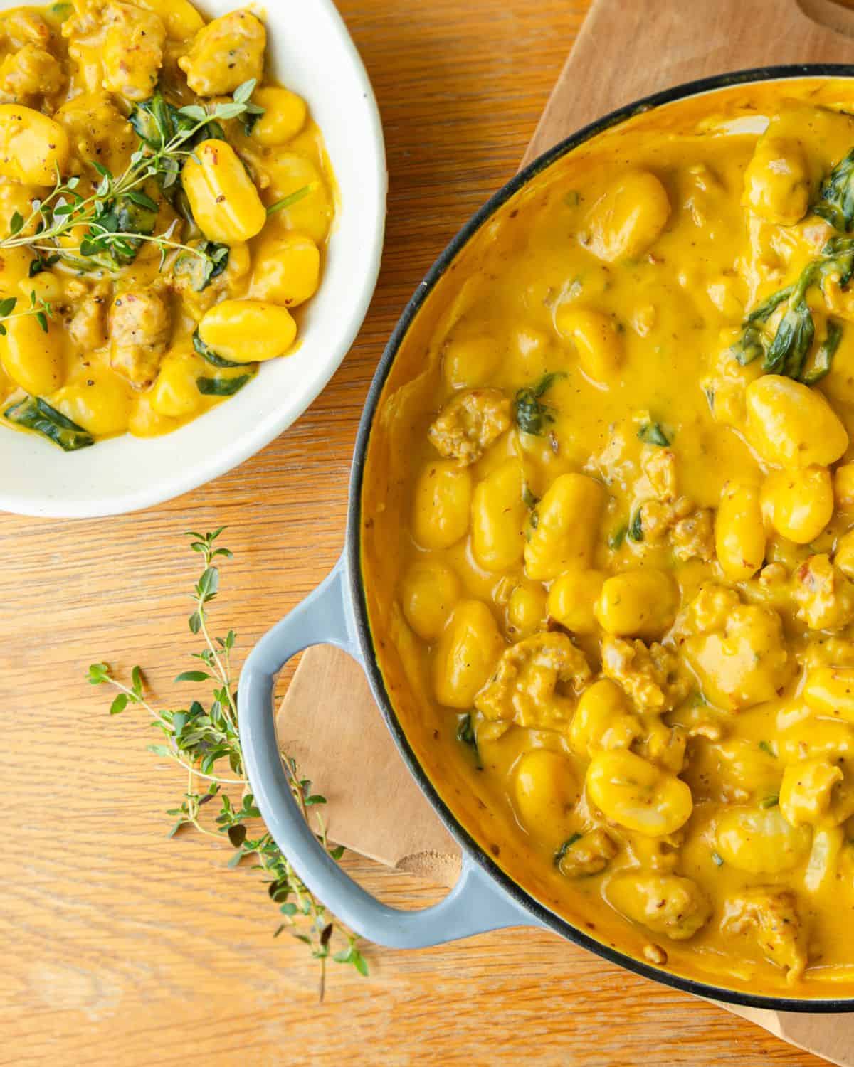 Creamy Pumpkin and Sausage Gnocchi A pot and a bowl of creamy gnocchi with spinach and sausage, garnished with fresh thyme, on a wooden surface.