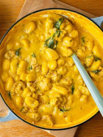 A pan filled with gnocchi in a creamy yellow sauce with spinach and chunks of sausage, sitting on a wooden surface.